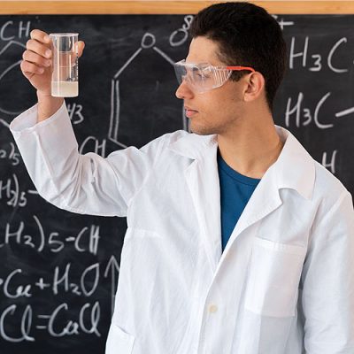 A male Arab chemistry teacher in goggles stands in a white coat against the background of a chalk board with formulas, holding a chemical flask with a reagent in his hands. copy space.